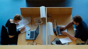 Studierende sitzen in der Bibliothek gegenüber an einem Schreibtisch an PCs mit Unterlagen.